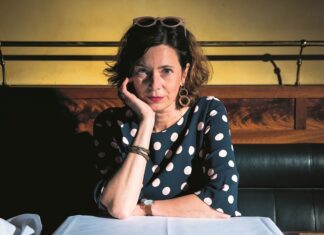 „Les Silences de Dunkelblum“ / Kulturforum Paris Das Bild zeigt die Autorin Eva Menasse sitzend auf einer dunklen Bank, mit einem Arm lehnend auf einem weiß gedeckten Tisch.