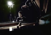 Alfredo Ovalles – Konzertreise / Kulturforum Tokio Das Bild zeigt den Pianisten Alfredo Ovalles vor schwarzem Hintergrund am geöffneten Flügel, hinter ihm ein Scheinwerfer, der sanftes Licht auf den Künstler und den geöffneten Flügel wirft. Die Spiegelung des Künstlers ist im geöffneten Flügel zu sehen.