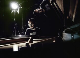 Alfredo Ovalles – Konzertreise / Kulturforum Tokio Das Bild zeigt den Pianisten Alfredo Ovalles vor schwarzem Hintergrund am geöffneten Flügel, hinter ihm ein Scheinwerfer, der sanftes Licht auf den Künstler und den geöffneten Flügel wirft. Die Spiegelung des Künstlers ist im geöffneten Flügel zu sehen.