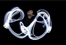 AN DIE FREUDE / White Hands Chorus Nippon & Mariko Tagashira Das Foto zeigt ein Kind vor einem dunklen Hintergrund, dessen Handbewegungen von Licht erhellt sind.