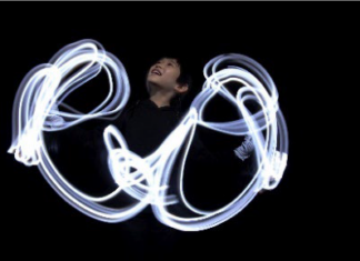 AN DIE FREUDE / White Hands Chorus Nippon & Mariko Tagashira Das Foto zeigt ein Kind vor einem dunklen Hintergrund, dessen Handbewegungen von Licht erhellt sind.