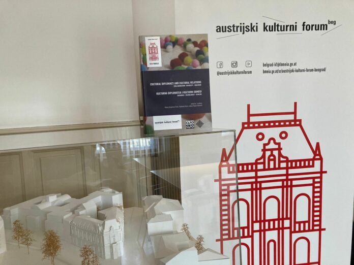 Das Bild zeigt gemeinsame AT-SRB Publikation zu Kulturdiplomatie vor KF Belgrad-Banner sowie Miniatur von ÖB/KF Belgrad und direkt gegenüber befindlicher Kunstuniversität Belgrad