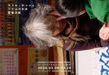 Anna Witt – Soft Skills, Thick Skin / Kulturforum Tokio Bild im Hochformat, gekippt. Zwei japanische Frauen fortgeschrittenen Alters, eine mit braunen Haaren, grünblauem Pullover und violettem Schal, sowie eine Frau mit grauen Haaren, bordeaux-rotem Pullover und orangem Schal. Beide Frauen haben ihre Augen geschlossen und werden von zwei Pflegekräften sanft im Gesicht berührt. Im Hintergrund findet sich eine orange Pinnwand mit einem Kalender und dem Foto einer Jugendfußballmanschaft.