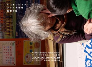 Anna Witt – Soft Skills, Thick Skin / Kulturforum Tokio Bild im Hochformat, gekippt. Zwei japanische Frauen fortgeschrittenen Alters, eine mit braunen Haaren, grünblauem Pullover und violettem Schal, sowie eine Frau mit grauen Haaren, bordeaux-rotem Pullover und orangem Schal. Beide Frauen haben ihre Augen geschlossen und werden von zwei Pflegekräften sanft im Gesicht berührt. Im Hintergrund findet sich eine orange Pinnwand mit einem Kalender und dem Foto einer Jugendfußballmanschaft.