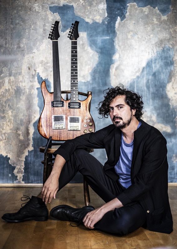 Das Foto zeigt den Musiker Saleh Rozati am Boden sitzend vor einer Wand mit einer doppelhalsigen Gitarre.