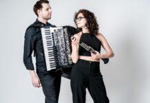 Duo Minerva / Kulturforum Warschau Das Bild zeigt zwei Personen vor einem hellen Hintergrund, eine Frau und einen Mann. Der Mann auf der linken Seite hält ein Akkordeon und trägt ein dunkles Hemd sowie eine dunkle Hose. Auf der rechten Seite steht eine Frau, die eine Klarinette spielt. Sie trägt ein schwarzes ärmelloses Oberteil und eine schwarze Hose. Beide Musiker scheinen sich auf eine musikalische Performance vorzubereiten. Der helle, einfarbige Hintergrund lenkt den Fokus auf die beiden Instrumente und ihre Leidenschaft für die Musik.