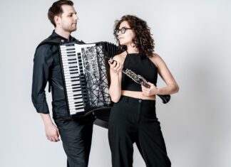 Duo Minerva / Kulturforum Warschau Das Bild zeigt zwei Personen vor einem hellen Hintergrund, eine Frau und einen Mann. Der Mann auf der linken Seite hält ein Akkordeon und trägt ein dunkles Hemd sowie eine dunkle Hose. Auf der rechten Seite steht eine Frau, die eine Klarinette spielt. Sie trägt ein schwarzes ärmelloses Oberteil und eine schwarze Hose. Beide Musiker scheinen sich auf eine musikalische Performance vorzubereiten. Der helle, einfarbige Hintergrund lenkt den Fokus auf die beiden Instrumente und ihre Leidenschaft für die Musik.