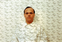 CineClub: Die Ameisenstraße / Kulturforum London Das Bild zeigt einen weißen Mann mit Ameisen-Hemd vor einer Tapete mit dem gleichen Ameisen-Muster