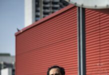 Das Ende ist nah / Kulturforum Bern Das Foto zeigt den Autor Amir Gudarzi stehend vor einer roten Häuserwand.