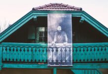 I, A TRANSPLANT // FROM OVERSEAS – W.H. AUDEN: KIRCHSTETTEN – NEW YORK / Kulturforum New York Das Bild zeigt die obere Hälfte eines Hauses mit Holzbalkon. Das Holz ist in türkiser Farbe gehalten. Am Dachgiebel ist ein Poster befestigt, dass den Dichter W.H. Auden mit Zigarette und Brille in der Hand zeigt.