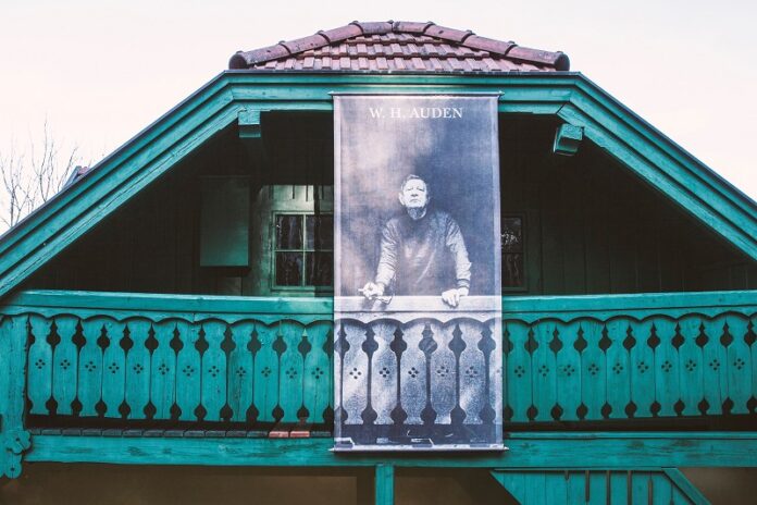 Das Bild zeigt die obere Hälfte eines Hauses mit Holzbalkon. Das Holz ist in türkiser Farbe gehalten. Am Dachgiebel ist ein Poster befestigt, dass den Dichter W.H. Auden mit Zigarette und Brille in der Hand zeigt.