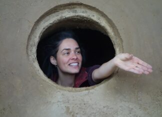 Calliope. Join the Dots / Botschaft Athen Auf dem Bild ist die Architektin und Hebamme Anka Duer zu sehen, sie streckt die Hand aus einer runden Öffnung - vermutlich aus dem auf Lehm gebauten Geburtsraum in unmittelbarer Nähe des Frauenmuseums Hittisau - entgegen.