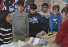 Film: Favoriten/ Generalkonsulat Hongkong Das Bild zeigt eine Lehrerin im linken Bildeck, die gerade sechs Schüler:Innen etwas erklärt. Dieser Film handelt von Kindern, die Deutsch nicht als Muttersprache haben. Sie stehen alle um einen Tisch und schauen die Lehrerin an.