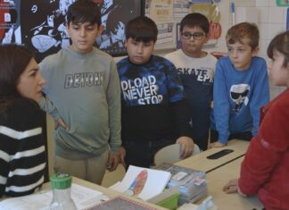 Film: Favoriten/ Generalkonsulat Hongkong Das Bild zeigt eine Lehrerin im linken Bildeck, die gerade sechs Schüler:Innen etwas erklärt. Dieser Film handelt von Kindern, die Deutsch nicht als Muttersprache haben. Sie stehen alle um einen Tisch und schauen die Lehrerin an.