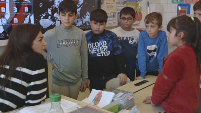 Das Bild zeigt eine Lehrerin im linken Bildeck, die gerade sechs Schüler:Innen etwas erklärt. Dieser Film handelt von Kindern, die Deutsch nicht als Muttersprache haben. Sie stehen alle um einen Tisch und schauen die Lehrerin an.