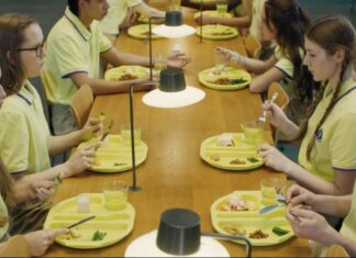 European Film Festival Hongkong / Generalkonsulat Hongkong Das Bild zeigt einen Mensatisch an dem sich Schüler in gelben Schuluniformen gegenübersitzen und essen. Es sind sehr kleine Portionen mit einem Glas Wasser und die Situation wirkt angespannt.