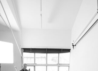 Merlin Ensemble Wien / Kulturforum Warschau Das Bild zeigt eine schwarz-weiße Aufnahme von drei Personen in einem hellen Raum mit hohen Decken und einem großen Fenster. Eine Person steht auf einer Leiter und hält eine Geige, während eine andere Person am Boden steht und ein Violoncello hält. Eine dritte Person steht neben ihnen, gegen die Wand mit dem Rücken angelehnt. Der Boden hat ein Holzmuster.