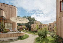 Ausstellung und Fachkonferenz / Kulturforum Bratislava Das Bild zeigt mehrere Gebäude aus Holz verbunden durch eine Grünfläche mit Weg. Auf dem haus vorne ist eine Terrasse mit Sonnenschirm und Liegen zu sehen.