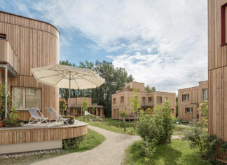 Ausstellung und Fachkonferenz / Kulturforum Bratislava Das Bild zeigt mehrere Gebäude aus Holz verbunden durch eine Grünfläche mit Weg. Auf dem haus vorne ist eine Terrasse mit Sonnenschirm und Liegen zu sehen.
