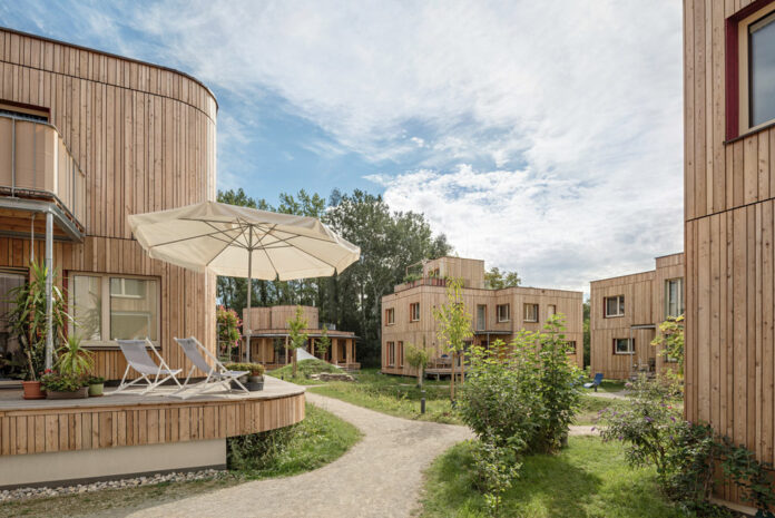 Das Bild zeigt mehrere Gebäude aus Holz verbunden durch eine Grünfläche mit Weg. Auf dem haus vorne ist eine Terrasse mit Sonnenschirm und Liegen zu sehen.