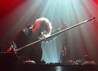 Mia Zabelka „PhotoTone“ / Kulturforum Brüssel Das Bild zeigt eine Frau mit kurzen Haaren, die vor einem Mischpult steht. Ihr Gesicht und der größte Teil ihres Körpers sind unkenntlich gemacht. In ihrer rechten Hand hält sie einen Geigenbogen. Von oben wird sie von einem Scheinwerfer angestrahlt, der den Scheitel ihres Haares hellblau beleuchtet.