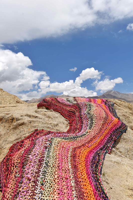 Das Bild zeigt das Kunstwerk “River of Sweat” von Anshu Singh, einen bunten gestrickten Teppich der über mehrere Wüstendünen verläuft