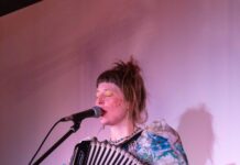 Konzert / Österreich-Bibliothek Vilnius Das Bild zeigt die Sängerin Alicia Edelweiss, die gleichzeitig in ein Mikrofon singt und ein Akkordeon spielt. Sie trägt ein buntes Kleid mit Blumen, eine blau-gelbe Kette, gelb-golden Lidschatten, rote Punkt auf den Wangen und roten Lippenstift.