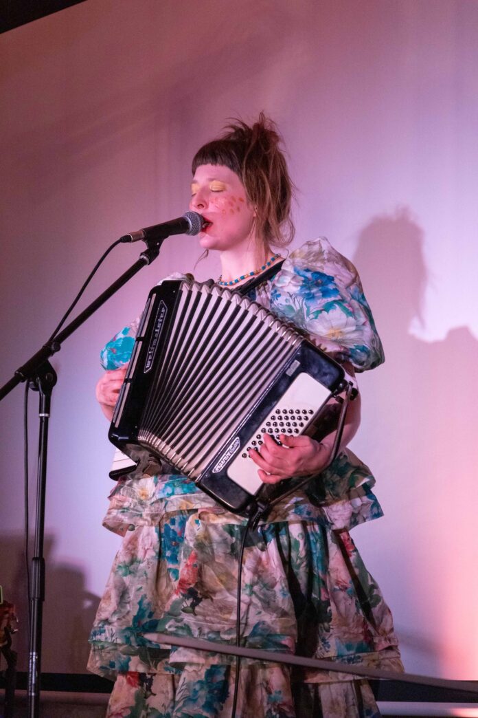 Das Bild zeigt die Sängerin Alicia Edelweiss, die gleichzeitig in ein Mikrofon singt und ein Akkordeon spielt. Sie trägt ein buntes Kleid mit Blumen, eine blau-gelbe Kette, gelb-golden Lidschatten, rote Punkt auf den Wangen und roten Lippenstift.