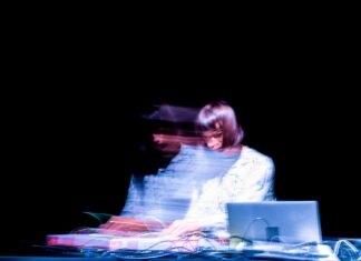 Konzert in Saskatoon/ Kulturforum Ottawa Das Bild zeigt die Künstlerin Isabella Forciniti. Sie spielt auf ihrem DJ-Set vor einem dunklen Hintergrund. Das Spotlight beleuchtet sie nur im Moment der Bewegung, während sie ihre Musik spielt. Sie trägt weiß und ihr Gesicht ist aufgrund der Bewegung nicht sichtbar.