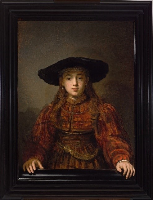 Das Bild zeigt ein Gemälde des Barockmalers Rembrandt. Das Gemälde bildet ein Mädchen mit Hut ab, das aus einem Bilderrahmen herausschaut bzw. herausgreift.