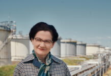 Margarethe Ottillinger – Theater in deutscher Sprache / Kulturforum Rom Das Bild zeigt die Schauspielerin Anita Zieher, verkleidet als Margarethe Ottillinger, als selbstbewusste Vorstandsdirektorin in Mantel, mit Kurzhaarschnitt, Brille und mit Aktentasche. Sie lächelt leicht und blickt in die Kamera. Im Hintergrund befindet sich ein Gelände, welches man einem Erdölkonzern zuordnen könnte - einem Bereich, mit dem Ottillinger in ihrer beruflichen Karriere über viele Jahre zu tun hatte.