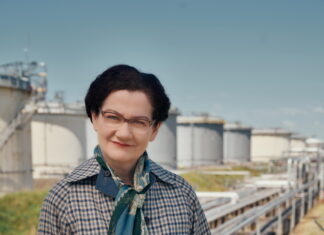 Margarethe Ottillinger – Theater in deutscher Sprache / Kulturforum Rom Das Bild zeigt die Schauspielerin Anita Zieher, verkleidet als Margarethe Ottillinger, als selbstbewusste Vorstandsdirektorin in Mantel, mit Kurzhaarschnitt, Brille und mit Aktentasche. Sie lächelt leicht und blickt in die Kamera. Im Hintergrund befindet sich ein Gelände, welches man einem Erdölkonzern zuordnen könnte - einem Bereich, mit dem Ottillinger in ihrer beruflichen Karriere über viele Jahre zu tun hatte.