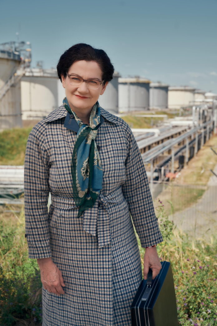 Das Bild zeigt die Schauspielerin Anita Zieher, verkleidet als Margarethe Ottillinger, als selbstbewusste Vorstandsdirektorin in Mantel, mit Kurzhaarschnitt, Brille und mit Aktentasche. Sie lächelt leicht und blickt in die Kamera. Im Hintergrund befindet sich ein Gelände, welches man einem Erdölkonzern zuordnen könnte - einem Bereich, mit dem Ottillinger in ihrer beruflichen Karriere über viele Jahre zu tun hatte.