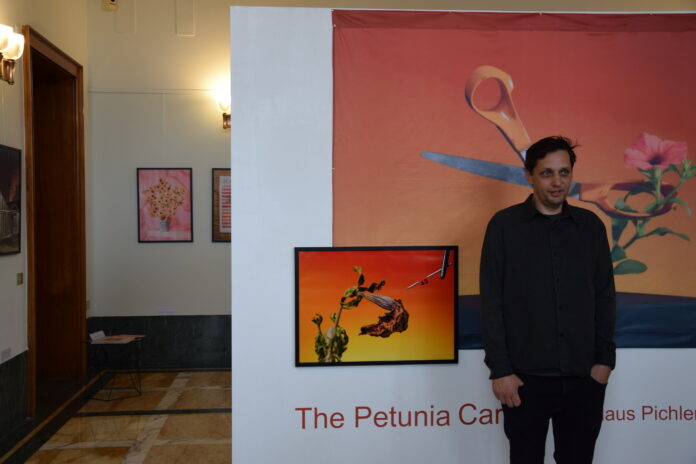 Das Bild zeigt den Fotograf Klaus Pichler bei der Ausstellungseröffnung von „THE PETUNIA CARNAGE“ im Atrium vom Österreichischen Kulturforum Rom. Er befindet sich vor einem seiner Werke, worauf man eine Schere sehen kann, welche eine orange, genmanipulierte Petuniensorte umschlingt und dabei auf ihre weltweite Zerstörung hingewiesen wird.