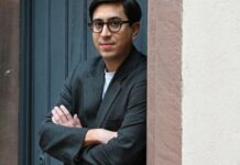 Tonio Schachinger in Ljubljana / Kulturforum Laibach Das Bild zeigt den Autor Tonio Schachinger an einer Tür lehnend mit verschränkten Armen,. Er trägt ein graues Sakko und eine schwarze Brille.