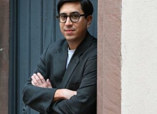 Tonio Schachinger in Ljubljana / Kulturforum Laibach Das Bild zeigt den Autor Tonio Schachinger an einer Tür lehnend mit verschränkten Armen,. Er trägt ein graues Sakko und eine schwarze Brille.