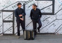 Folkoteque: Oscar Antolí & Milos Todorovski / Kulturforum Brüssel Das Bild zeigt zwei Männer, die vor einer weißen Backsteinmauer stehen. Die Wand ist mit schwarzen Streifen beschmiert, die an übereinander liegende Balken erinnern. Beide Männer tragen ein schwarzes Hemd, eine schwarze Hose und schwarze Turnschuhe. Der Mann auf der linken Seite hat kurzes gelocktes braunes Haar und einen Vollbart. Seine Hände schweben wie in Bewegung über seiner Brust, sein Gesicht ist nach unten geneigt und seine Hüfte ist zur Seite geneigt, wobei sein rechtes Bein weiter herausragt als das linke. Der andere Mann, der auf der rechten Seite des Bildes zu sehen ist, blickt den anderen Mann an und lächelt. Seine Hände stecken in den Hosentaschen, sein hellbraunes Haar ist zu einem tiefen Dutt zusammengefasst. Sein linker Fuß lehnt mit dem Rücken an der Wand. Im vorderen Teil des Bildes, der die Beine des Mannes teilweise verdeckt, ist ein Akkordeon zu sehen.