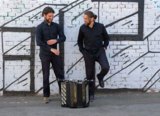 Folkoteque: Oscar Antolí & Milos Todorovski / Kulturforum Brüssel Das Bild zeigt zwei Männer, die vor einer weißen Backsteinmauer stehen. Die Wand ist mit schwarzen Streifen beschmiert, die an übereinander liegende Balken erinnern. Beide Männer tragen ein schwarzes Hemd, eine schwarze Hose und schwarze Turnschuhe. Der Mann auf der linken Seite hat kurzes gelocktes braunes Haar und einen Vollbart. Seine Hände schweben wie in Bewegung über seiner Brust, sein Gesicht ist nach unten geneigt und seine Hüfte ist zur Seite geneigt, wobei sein rechtes Bein weiter herausragt als das linke. Der andere Mann, der auf der rechten Seite des Bildes zu sehen ist, blickt den anderen Mann an und lächelt. Seine Hände stecken in den Hosentaschen, sein hellbraunes Haar ist zu einem tiefen Dutt zusammengefasst. Sein linker Fuß lehnt mit dem Rücken an der Wand. Im vorderen Teil des Bildes, der die Beine des Mannes teilweise verdeckt, ist ein Akkordeon zu sehen.