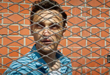 Sommerkino Lyon – Die Große Freiheit (Sebastian Meise) / Kulturforum Paris Das Bild zeigt den Schauspieler Franz Rogowski in blauem Hemd vor einer roten Ziegelmauer und hinter einem Gitterzaun