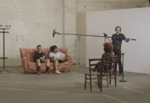 MUTZENBACHER in Prag / Kulturforum Prag Das Bild zeigt eine Filmszene mit zwei Männern auf einem rosafarbenen Sofa. Gegenüber von ihnen sitzt die Regisseurin Ruth Beckermann, und daneben steht ein Mann mit Technik