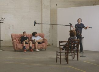 MUTZENBACHER in Prag / Kulturforum Prag Das Bild zeigt eine Filmszene mit zwei Männern auf einem rosafarbenen Sofa. Gegenüber von ihnen sitzt die Regisseurin Ruth Beckermann, und daneben steht ein Mann mit Technik