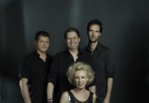 Konzert Michaela Rabitsch & Robert Pawlik / Kulturforum Warschau Auf dem Bild sind vier Personen zu sehen. Im Vordergrund sitzt eine Frau auf einem Hocker. Sie hat kurze, blonde, lockige Haare und trägt eine glänzende, ärmellose schwarze Bluse sowie schwarze Lederhosen. Sie lächelt in die Kamera. Hinter ihr stehen drei Männer. Alle sind dunkel gekleidet. Der Mann links trägt ein schwarzes Hemd und blickt ebenfalls in die Kamera. Der Mann in der Mitte hat schulterlange braune Haare, lächelt und trägt ein dunkles Hemd. Der Mann rechts hat kurzes Haar und trägt ein schwarzes Hemd. Auch er blickt in die Kamera. Der Hintergrund des Bildes ist dunkel, was die Personen im Vordergrund hervorhebt. Das Licht ist so gesetzt, dass die Gesichter und Oberkörper der Personen gut ausgeleuchtet sind.