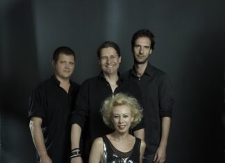 Konzert Michaela Rabitsch & Robert Pawlik / Kulturforum Warschau Auf dem Bild sind vier Personen zu sehen. Im Vordergrund sitzt eine Frau auf einem Hocker. Sie hat kurze, blonde, lockige Haare und trägt eine glänzende, ärmellose schwarze Bluse sowie schwarze Lederhosen. Sie lächelt in die Kamera. Hinter ihr stehen drei Männer. Alle sind dunkel gekleidet. Der Mann links trägt ein schwarzes Hemd und blickt ebenfalls in die Kamera. Der Mann in der Mitte hat schulterlange braune Haare, lächelt und trägt ein dunkles Hemd. Der Mann rechts hat kurzes Haar und trägt ein schwarzes Hemd. Auch er blickt in die Kamera. Der Hintergrund des Bildes ist dunkel, was die Personen im Vordergrund hervorhebt. Das Licht ist so gesetzt, dass die Gesichter und Oberkörper der Personen gut ausgeleuchtet sind.