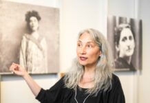 Pride Month / Kulturforum Warschau Das Foto zeigt Petra Unger, die eine schwarze Bluse und eine silberne Halskette trägt. Petra erzählt gerade etwas. Hinter ihr, im Hintergrund, sind unscharfe Bilder von Frauen an der Wand zu sehen.