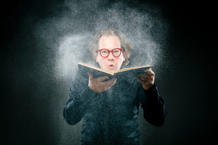 Das Bild zeigt Norbert Trawöger mit roter Brille mit einem Buch, von dem er Staub wegbläst.