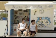 CineClub: Vista Mare / Kulturforum London Das Bild zeigt zwei sommerlich bekleidete junge Männer vor einem reichlich beschmückten Strandhaus. Beide schauen durch die Sonnenbrille nach rechts