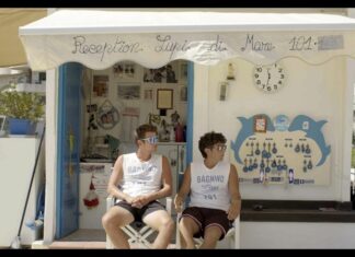 CineClub: Vista Mare / Kulturforum London Das Bild zeigt zwei sommerlich bekleidete junge Männer vor einem reichlich beschmückten Strandhaus. Beide schauen durch die Sonnenbrille nach rechts