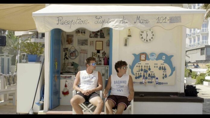 Das Bild zeigt zwei sommerlich bekleidete junge Männer vor einem reichlich beschmückten Strandhaus. Beide schauen durch die Sonnenbrille nach rechts