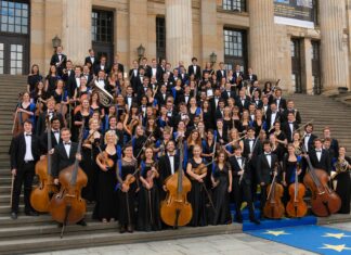 European Union Youth Orchestra/ Kulturforum New York Das Bild zeigt das European Union Youth Orchestra. Die Orchestermitglieder sind alle in schwarz-weiß gekleidet und halten – wo möglich – ihre Instrumente in Händen. Sie tragen zusätzlich einen blauen dünnen Schal