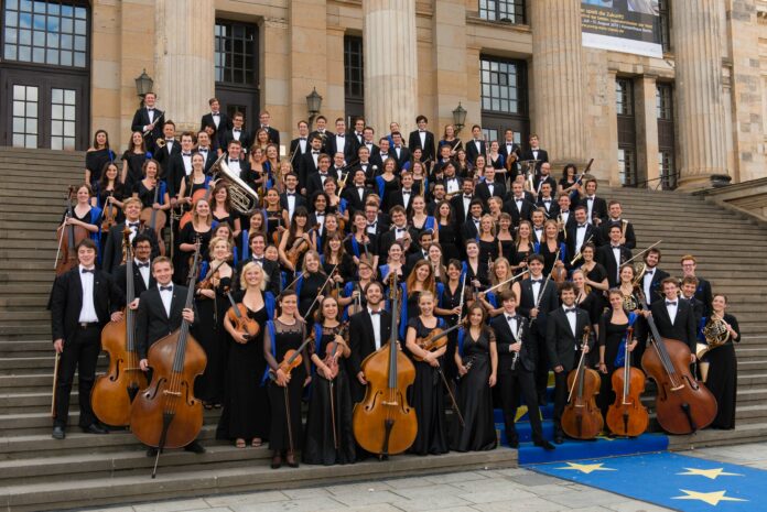 Das Bild zeigt das European Union Youth Orchestra. Die Orchestermitglieder sind alle in schwarz-weiß gekleidet und halten – wo möglich – ihre Instrumente in Händen. Sie tragen zusätzlich einen blauen dünnen Schal